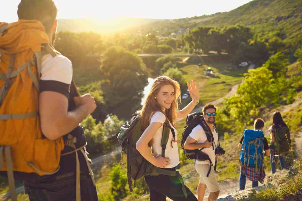 Groupe de personnes en randonné avec une femme blonde avec une t-shirt blanc et un sac à dos sourit à la caméra à côté d'un homme brun en t-shirt blanc avec des lunettes de soleil